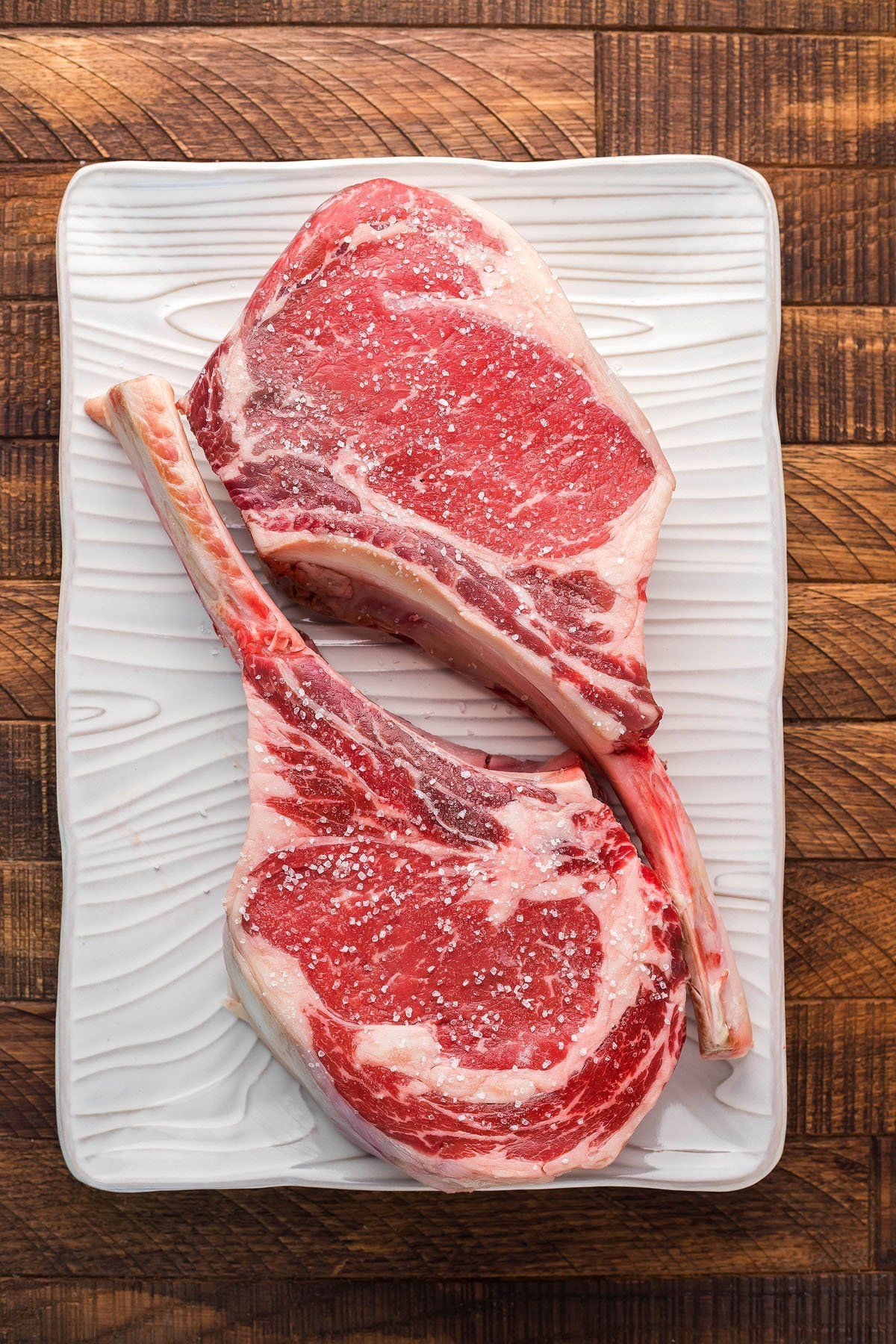 Two grass-fed tomahawk ribeyes, salted, resting on a white platter
